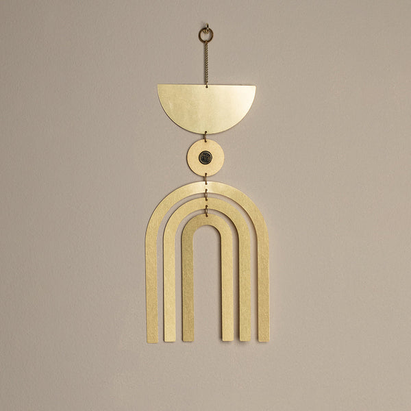 The Supernova Pyrite Wall Hanging by Scout Jewelry is a minimalist piece made from gold-toned metal, featuring a semicircular top, a small central circle with a semi-precious stone, and three downward arch shapes. It is elegantly displayed against a light beige background.