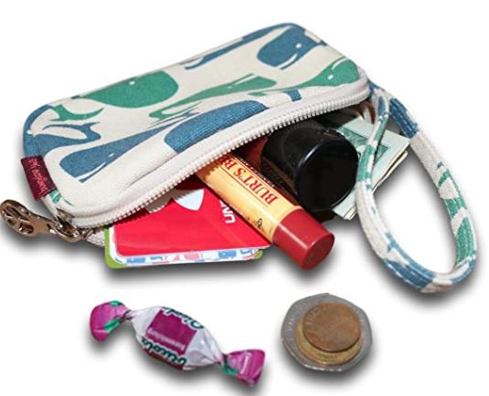 The Tiny Wristlet Whale by Bungalow 360, made from natural cotton canvas, is open to reveal its contents: a tube of Burt's Bees lip balm, a red card, a small bottle, and a key. In front of the wristlet are wrapped candy and some coins. This vegan clutch features an eye-catching whale design and includes a wrist strap for convenience.