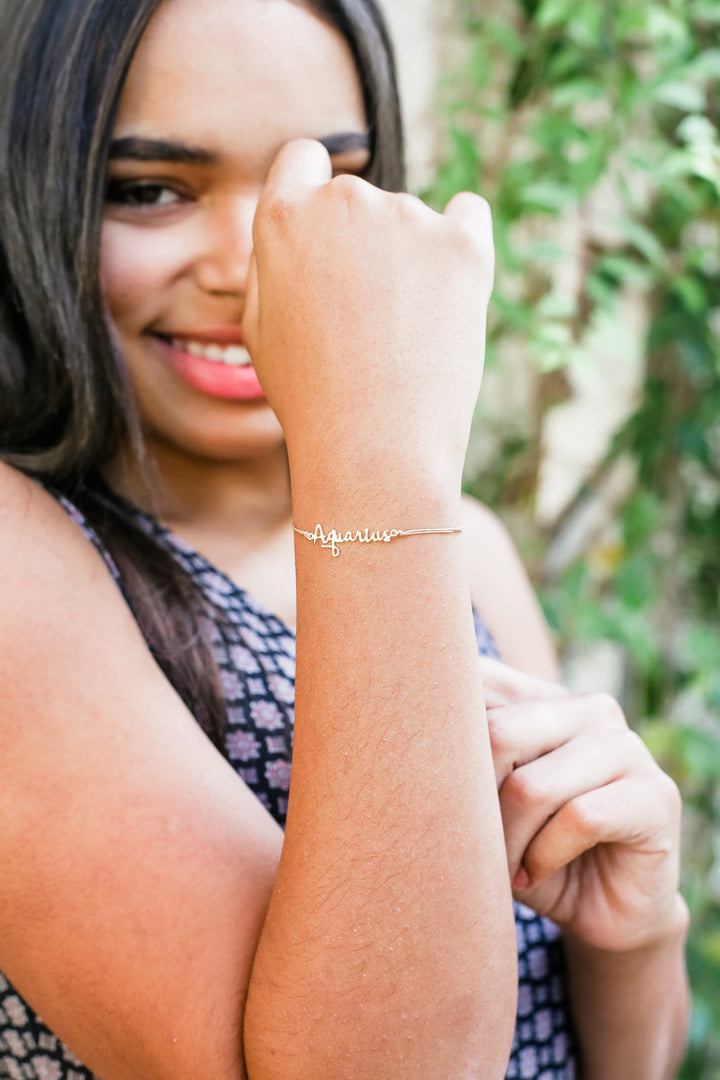 A person smiles while showcasing a zodiac bracelet on their wrist with "Aries" elegantly displayed. Their long hair cascades over a patterned top, and the 14k gold-dipped finish of the LuckyFeather bracelet glimmers softly against the blurred greenery backdrop.