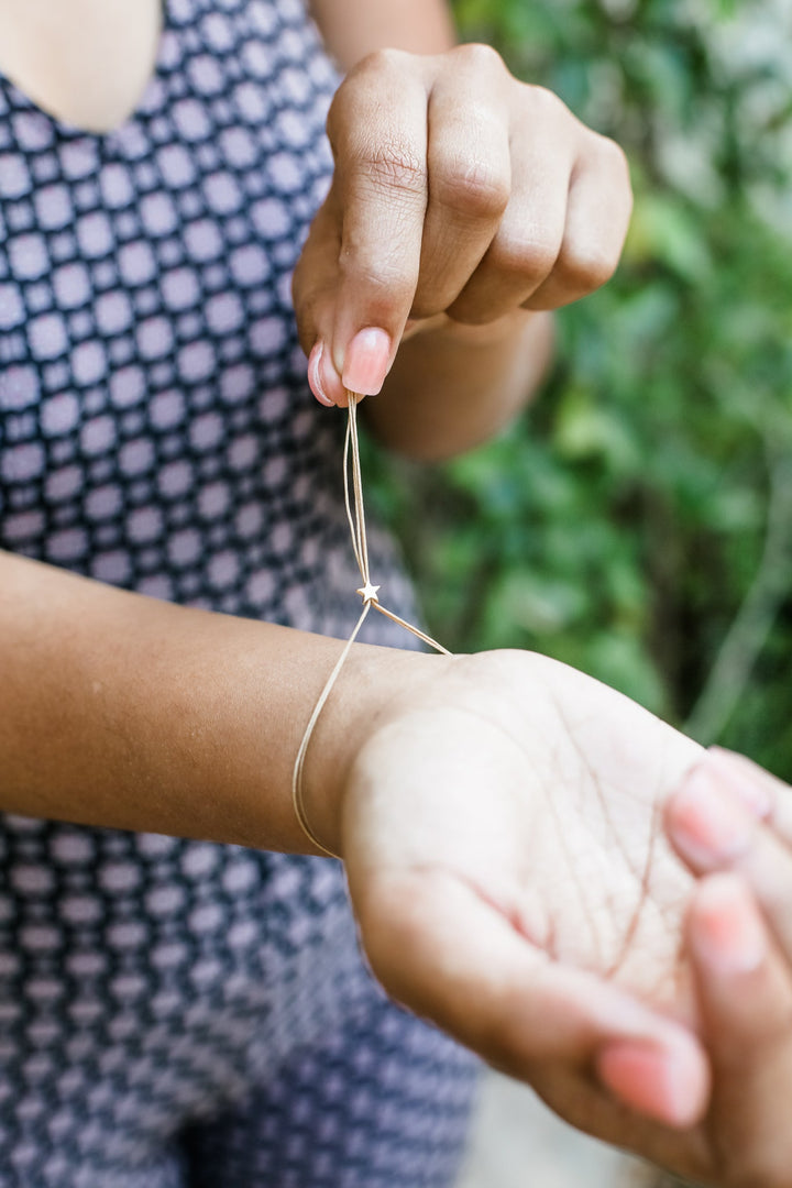Close-up of someone wearing a patterned sleeveless top, tying an adjustable Zodiac Bracelet Leo by LuckyFeather around their wrist. The other hand secures the loop. Blurred greenery in the background perfectly complements the delicate charm of the Zodiac collection.