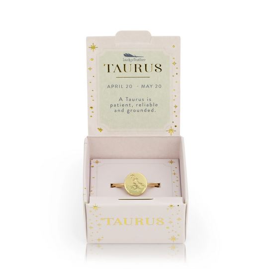 A 14k gold-dipped Taurus Signet Ring by LuckyFeather is presented in an open pink box. The lid reads "TAURUS, APRIL 20 - MAY 20. A Taurus is patient, reliable and grounded" with stars decorating the inside. "TAURUS" and stars adorn the front of the box, which features an adjustable band for a perfect fit.