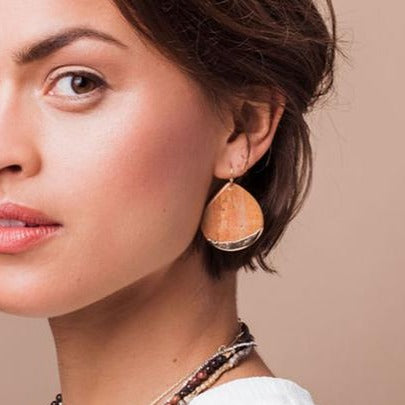 A person with short hair showcases a stunning, large Scout Jewelry 'Dipped Teardrop Earring' made from gold-dipped metal and petrified wood, complemented by layered necklaces. The solid tan background enhances their look. They're dressed in a white top, with only the left side of their face visible in the close-up.