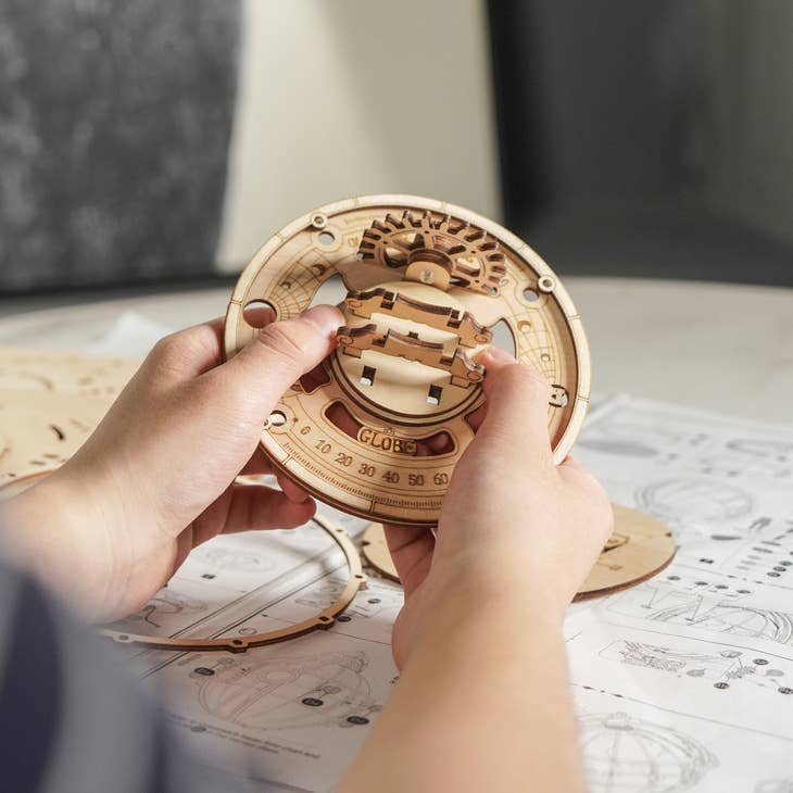 Hands assembling the DIY Wooden Puzzle: Luminous Globe by Hands Craft. The puzzle piece features intricate gears and markings, with additional pieces and an instruction manual visible in the background.