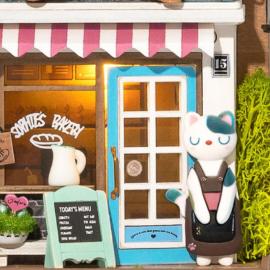 The Hands Craft DIY Miniature Wall Hanging Kit: Aroma Toast Lab features a cute cat figurine in an apron outside a bakery with real lighting, a striped awning, menu board, blue-framed entrance, and milk jug illustration through the window.