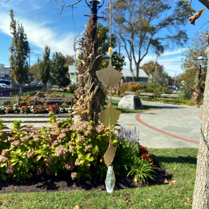 Beneath a vivid blue sky, vibrant flowers and trees create a picturesque garden scene. In the foreground, hanging elegantly from a tree branch, is the Leaf Amazonite Suncatcher by Scout Jewelry. This exquisite piece features leaf-shaped ornaments and a crystal drop that casts rainbows as it gracefully reflects sunlight.