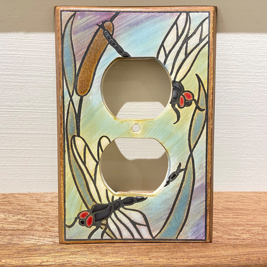 The Receptacle Dragonfly Bulrush by ALL FIRED UP is a decorative outlet cover adorned with painted dragonfly and red-eyed insect motifs on a ceramic switch plate. Made in Canada, it showcases a backdrop of gentle pastel hues, finished with transparent screws for a seamless appearance.