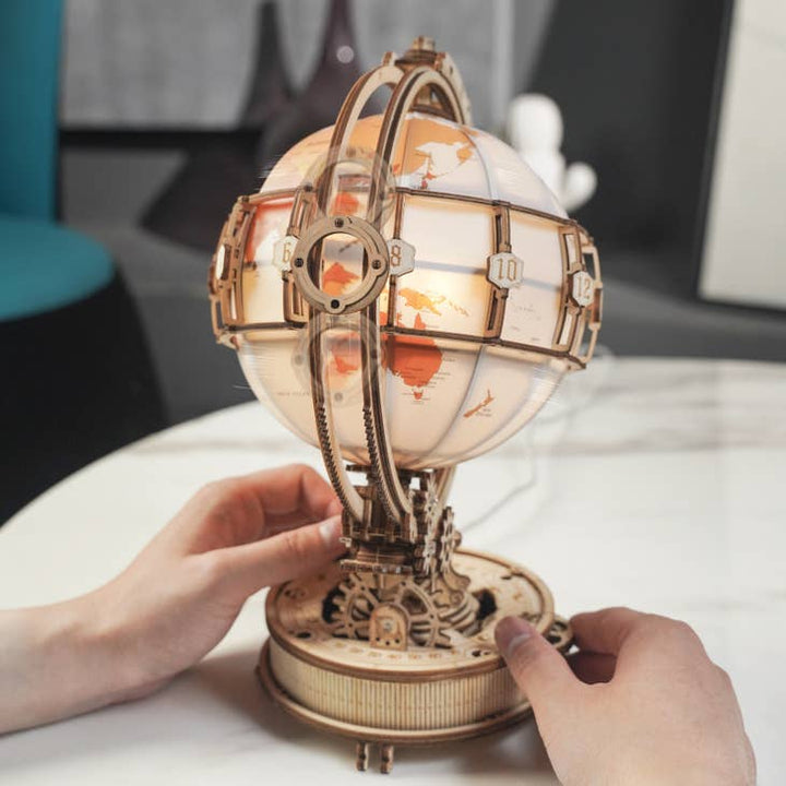 A person adjusts the Hands Craft DIY Wooden Puzzle: Luminous Globe on a table. The intricate design features gears and numbered plates around the globe, indicating it is a clock or timepiece. This 3D wooden puzzle showcases a blend of craftsmanship and engineering, doubling as a mesmerizing night light.