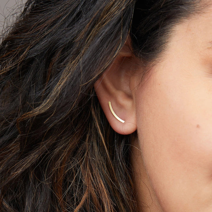 Close-up of a woman's ear adorned with the Comet Curve Gold earring by Scout Jewelry, an 18k gold vermeil piece shaped like a curved line. Her dark hair has lighter streaks, and the image emphasizes the earring's sleek and elegant minimalist design.