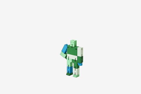 The Cubebot Micro Green Multi, a small, multicolored humanoid toy figure by Areaware, stands against a plain white background. Reminiscent of Japanese Shinto Kumi-ki puzzles, it is constructed from rectangular blocks in various shades of green and blue. The figure appears to be in a casual pose with its left arm slightly raised.