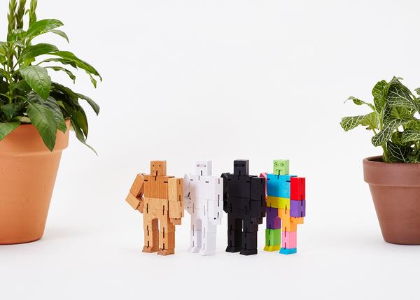 Four Areaware Cubebot Micro Purple Multi wooden toy robots, inspired by Japanese Shinto Kumi-ki puzzles, stand between two potted plants on a white background. Each figure showcases a different color: natural wood, white, black, and multicolored.