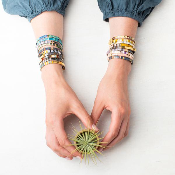 Hands adorned with Scout Jewelry's Miyuki Good As Gold Forest Gold bracelets elegantly hold a small green air plant above a white surface, while the person wears a long-sleeved blue top.