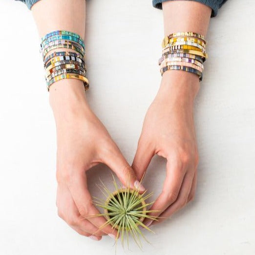 Two hands adorned with Scout Jewelry's Miyuki Pure Magic Gold bracelets gently hold a small green air plant on a white surface, forming a heart shape with their fingers. The person's forearms are positioned parallel, showcasing the empowering messages on the stack of bracelets.