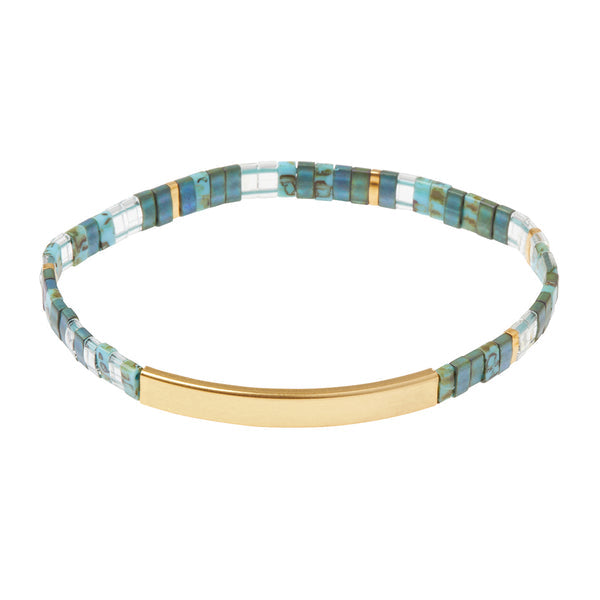 The Scout Jewelry "Miyuki Joy and Kindness Marine Gold" bracelet, showcasing minimalist design with Japanese glass Miyuki beads in blue and clear shades, is accented by a 14K gold dipped bar against a white background. This combination of colors and materials creates a modern and elegant look.