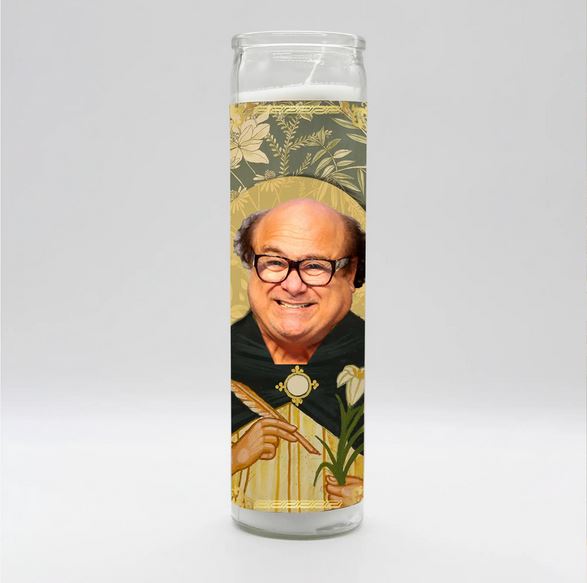 The Danny DeVito Candle from BOBBYK BOUTIQUE features a berry scent and a smiling depiction of Danny DeVito with glasses and a receding hairline, set against a decorative floral backdrop. He is portrayed in a saint-like image, holding a stem with flowers and wearing green and gold robes, illuminated by the gentle candlelight.