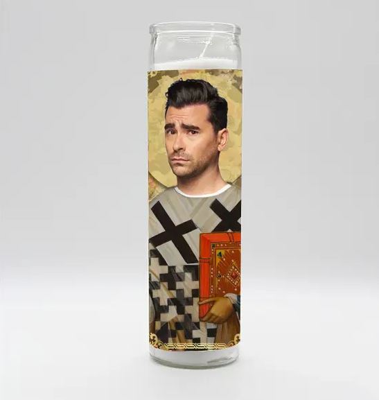 The David Rose Candle by BOBBYK BOUTIQUE showcases a stylized image of a man with dark hair and a serious expression, dressed in patterned clothing adorned with black crosses, and holding a red book. This tall white candle features a berry scent and boasts an ornate golden background design.