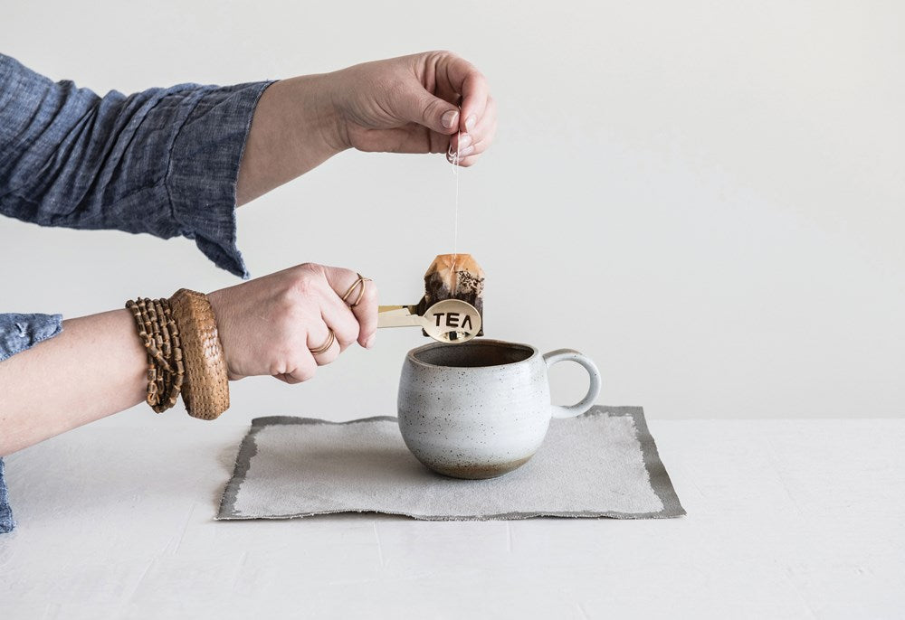 A tea enthusiast wearing a blue shirt and wooden bracelets uses CreativeCoOp's Stainless Steel Tea Tongs Gold to dip a tea bag labeled "TEA" into a speckled ceramic mug, which sits on a gray mat on top of a white table.