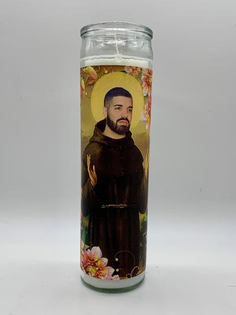 The Drake Candle by BOBBYK BOUTIQUE is a tall unscented candle featuring an illustrated bearded, shaved-head man in a saint-like pose, wearing a brown robe and halo. He is surrounded by flowers on a plain background, reminiscent of a celebrity shrine.