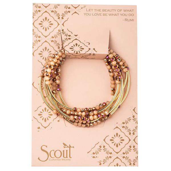 The Wrap Dune/Gold bracelet by Scout Jewelry is beautifully showcased on a decorative card, featuring a quote starting with "Let the beauty of what" adorned with ornate designs and the Scout brand logo. The multi-strand Scout Wrap bracelet elegantly combines wooden and crystal beads with gold-colored strands and purple accents, emphasizing its sophisticated allure.