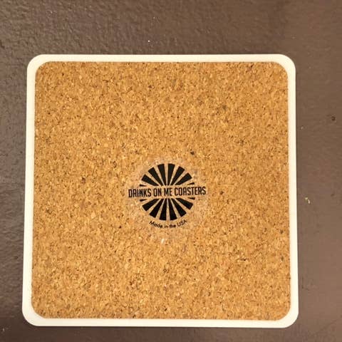 A top-down view showcases the Burning Sage Coaster from Drinks On Me. This cork coaster, framed with a stylish white border, features a black sunburst graphic at its center, along with the text "DRINKS ON ME COASTERS" and "Made in the USA" below it. The piece boasts a durable resin finish and cork backing to protect your surfaces.