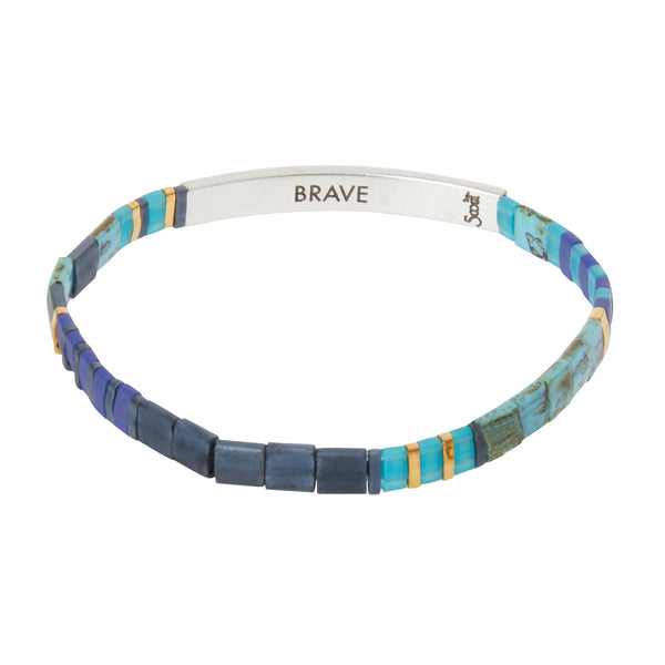 The Miyuki Brave Cobalt Silver bracelet from Scout Jewelry features a delicate band adorned with vibrant Japanese glass Miyuki beads in shades of blue, purple, and turquoise, interspersed with small gold accents. The metal clasp is engraved with the word "BRAVE" and a small tribal symbol, conveying empowering messages to the wearer.