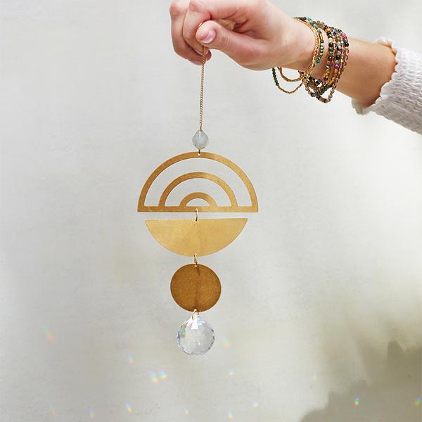 A hand, adorned with colorful beaded bracelets, holds the Scout Jewelry "Rainbow Labradorite Suncatcher." The suncatcher features an arc, a semi-circle, and two circular shapes with a faceted crystal at the bottom that acts as a rainbow maker, all connected by a chain. The background is a light, plain wall.