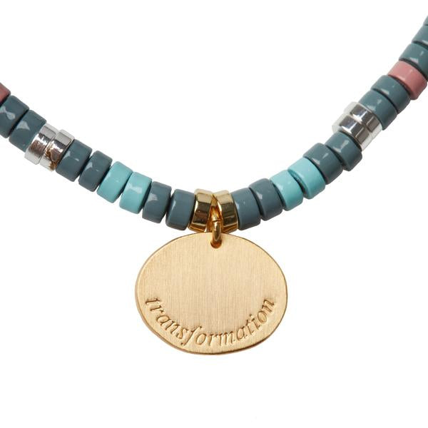 The Intention Charm African Turq/Gold necklace by Scout Jewelry features multi-colored bead strands in shades of teal, pink, gray, and silver. Its centerpiece is a gold pendant engraved with the word "transformation," enhanced by semi-precious stones that radiate elegance and positivity.