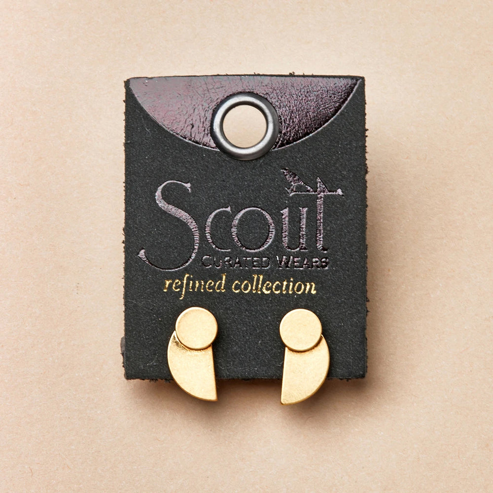 A pair of "Eclipse Stud Gold" earrings by Scout Jewelry, made of 18k gold vermeil and shaped like commas, displayed on a dark green card with the text "Scout Curated Wears Refined Earring Collection" printed on it. The card features a circular hole at the top for hanging against a light beige background.