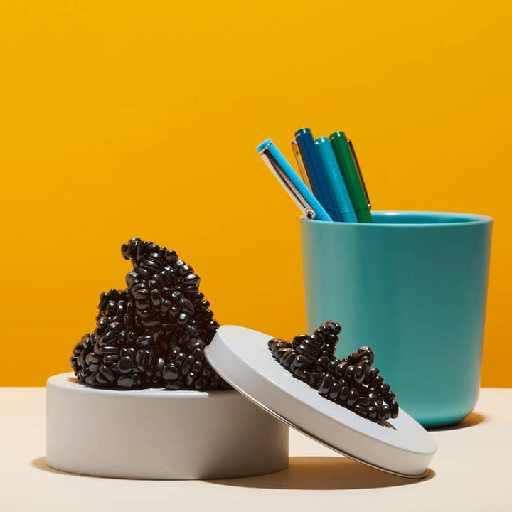 A container of Crags Magnetic Putty Stones by SPEKS, shaped like glossy black beans with its lid off, sits beside a teal pen cup against a bright orange background, evoking stress relief.