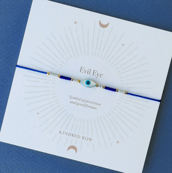 The Evil Eye Cord Bracelet Blue, featuring a small evil eye charm and a thin blue cord, is displayed on a white card with a radial design and the text "Evil Eye. Symbol of protection against bad luck and good fortune." The card bears the Kindred Row branding.