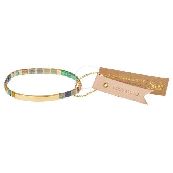 The Miyuki Good As Gold Forest Gold bracelet by Scout Jewelry features multicolored square beads, a 14K gold-dipped bar, and a paper tag with "Good Karma Bracelet" and "Good As Gold." Crafted on a stretch cord with a small gold chain, it complements the Japanese glass Miyuki bracelets collection.
