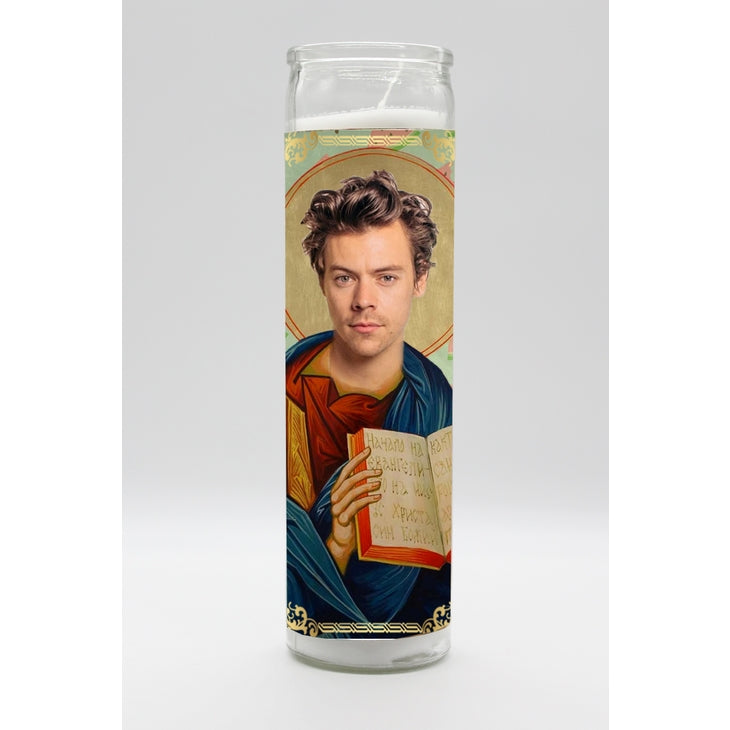 The Harry Styles Candle from BOBBYK BOUTIQUE showcases a portrait of a young man with styled hair, dressed in traditional robes and holding an open book. The design mimics religious iconography with a halo around his head. Hand-poured and featuring a light gray background, this tall white candle emits a subtle berry scent when lit.