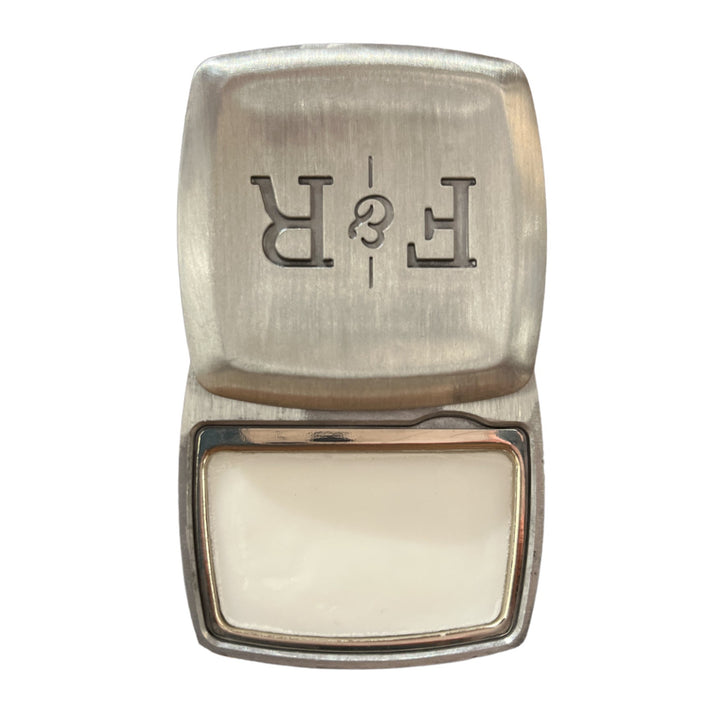 A brushed metallic rectangular locket, partially open to reveal a white, glossy surface inside. The top features an engraved mirrored "R & E" and carries a faint sandalwood scent. This is the Calle Ocho .2oz by Fulton & Roark.