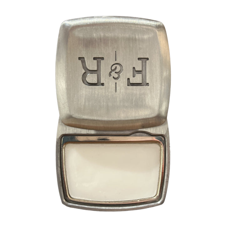 A silver metal stamp with the letters "Fulton & Roark" etched in reverse on the top part, reminiscent of the unique beauty at Green Sand Beach. The stamp is open to reveal a white rectangular pad inside, evoking the essence of Mahana .2oz's solid fragrance.