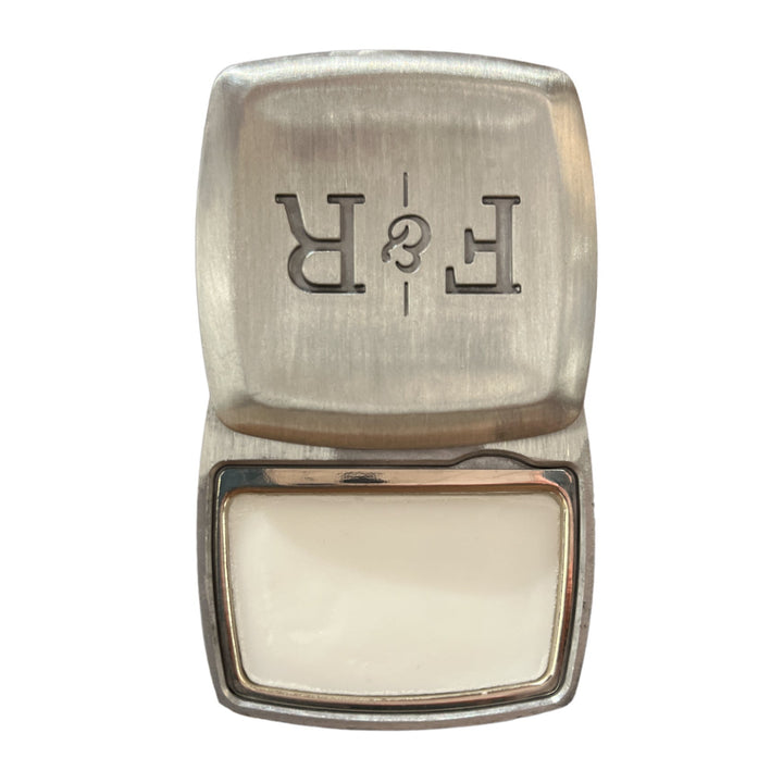 A metal container with a hinged lid showcases the letters "R & E" in reverse. The open lid reveals the white, vanilla-scented balm inside. Sterling .2oz, by Fulton & Roark, features a brushed metal finish that feels as sleek as leather.