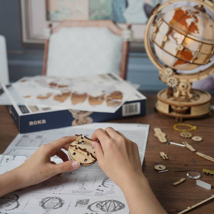 A person is assembling the Hands Craft DIY Wooden Puzzle: Luminous Globe using detailed instructions. Various parts and tools are scattered on the table, and a partially assembled globe is visible. In the background, an opened box with more pieces is on the chair, hinting at a creative night light project in progress.