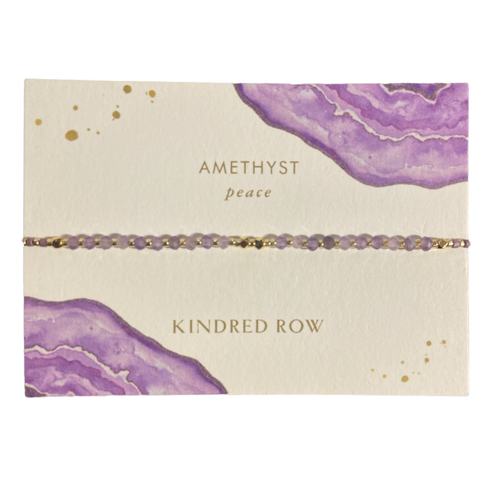 The Amethyst Healing Gemstone Stacking Bracelet by Kindred Row is a delicate piece featuring small amethyst beads. It is elegantly displayed on a card with purple watercolor designs, with the inscriptions "Amethyst peace" and "Kindred Row." This handmade bracelet showcases natural gemstones on an adjustable cord.