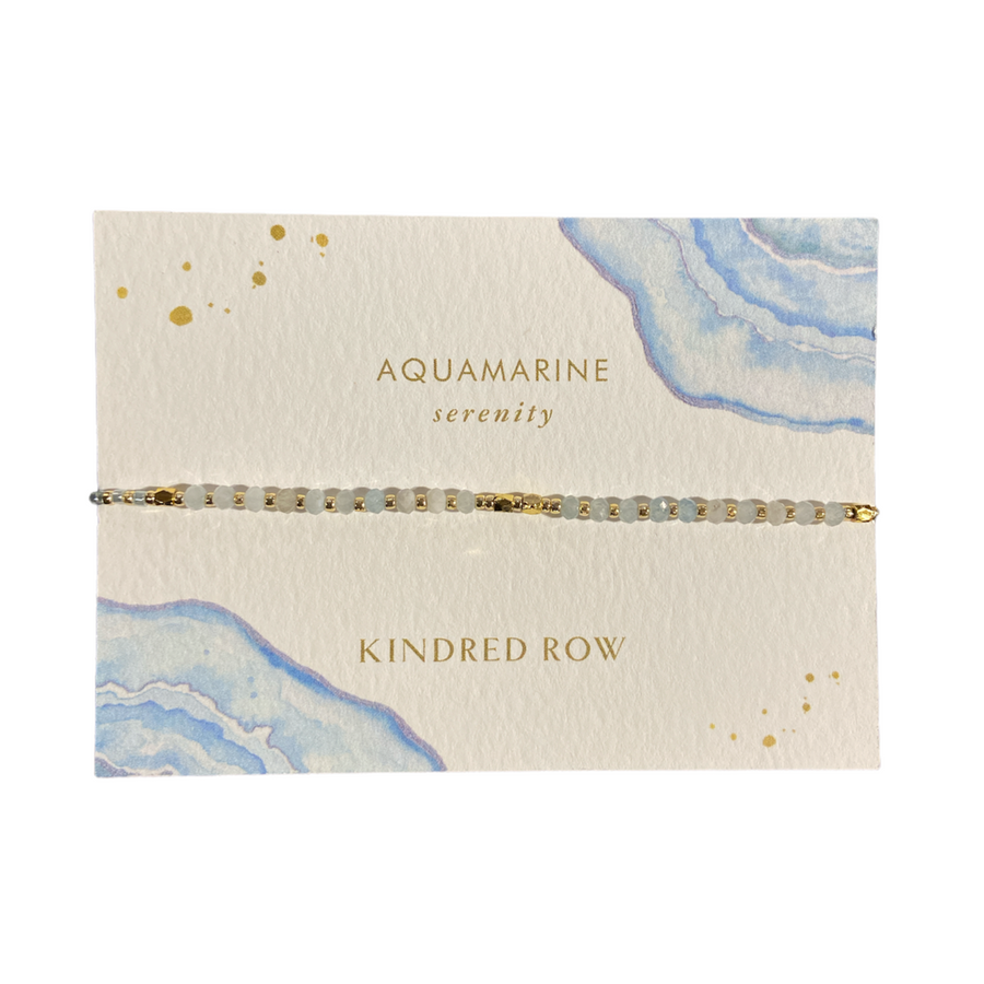 The Aquamarine Healing Gemstone Stacking Bracelet from Kindred Row, made with natural gemstones, is elegantly showcased on a decorative card featuring a watercolor blue background and gold accents. This handmade piece rests on an adjustable soft cotton cord. The card displays "AQUAMARINE serenity" and "KINDRED ROW" in gold lettering.