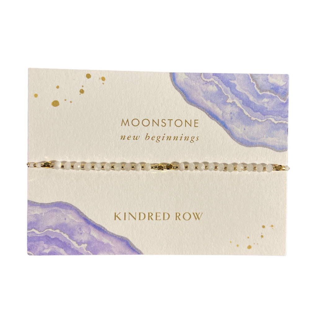 A Moonstone Healing Gemstone Stacking Bracelet featuring an adjustable cord is elegantly presented on a white card adorned with purple watercolor accents. "Moonstone, new beginnings" and "Kindred Row" are elegantly inscribed in gold lettering, emphasizing the charm of these natural gemstones.