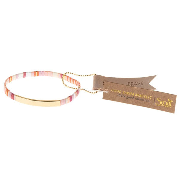 The Miyuki Brave Gold bracelet by Scout Jewelry is multicolored with pink, orange, and white beads, featuring a 14K gold dipped bar. Two brown tags read "BRAVE" and "GOOD KARMA BRACELET," offering empowering messages for the wearer.