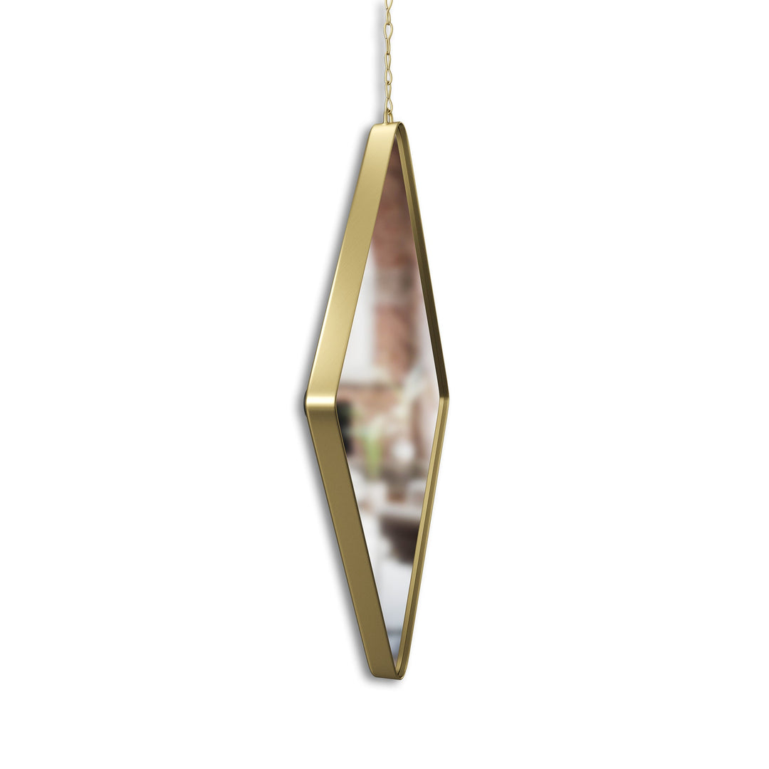 The Umbra Diamond Mirror Brass hangs against a white background, adding an elegant touch. Its blurred surface offers a distorted reflection of an unidentifiable setting.