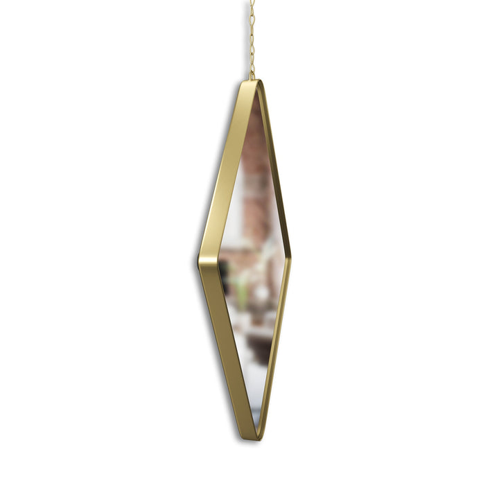 The Umbra Diamond Mirror Brass hangs against a white background, adding an elegant touch. Its blurred surface offers a distorted reflection of an unidentifiable setting.