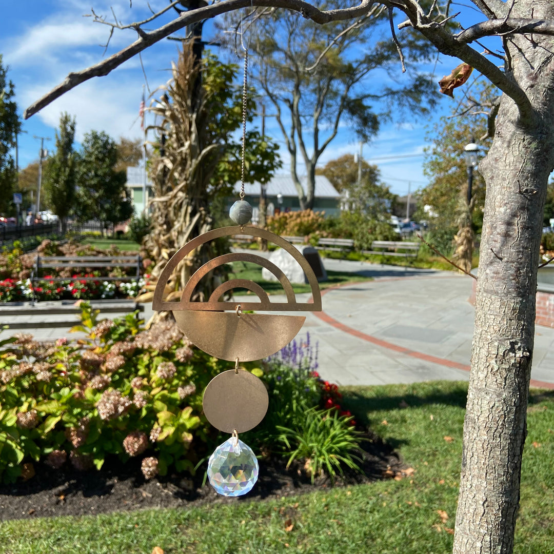 A close-up of the Rainbow Labradorite Suncatcher by Scout Jewelry hanging from a tree branch in a park. The suncatcher features a geometric design with a crescent and circular pendant, ending with a clear crystal ornament that acts as a rainbow maker. The background shows flowers, greenery, and a stone pathway under a clear blue sky.