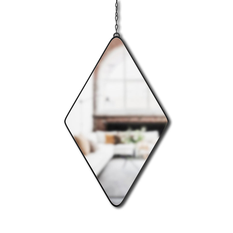 An Umbra Diamond Mirror Black, with its diamond-shaped design and button-like quality, hangs from a chain, reflecting a blurred view of a cozy, sunlit living room featuring a white sofa, pillows, and a coffee table. Large windows let in natural light, enhancing the soft and inviting ambiance.