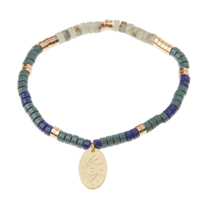 Introducing the Intention Charm Labra/Gold by Scout Jewelry: a beaded bracelet adorned with an array of small, cylindrical beads in lush shades of green, blue, and white, highlighted with metallic gold accents. Featuring a 14K gold dipped charm pendant engraved with an eye symbol that dangles elegantly from the bracelet, this piece showcases semi-precious stones celebrated for their positive properties.