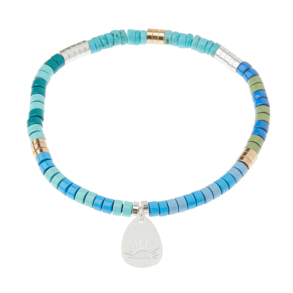 Introducing the Intention Charm Turq/Silv/Gold by Scout Jewelry - a beaded bracelet featuring shades of blue, green, and silver semi-precious stones. The uniformly sized cylindrical beads create a harmonious look, complemented by a small metallic charm dipped in 14K gold and engraved with a stylized sun that hangs elegantly from the bracelet. The background is white, enhancing its striking design.