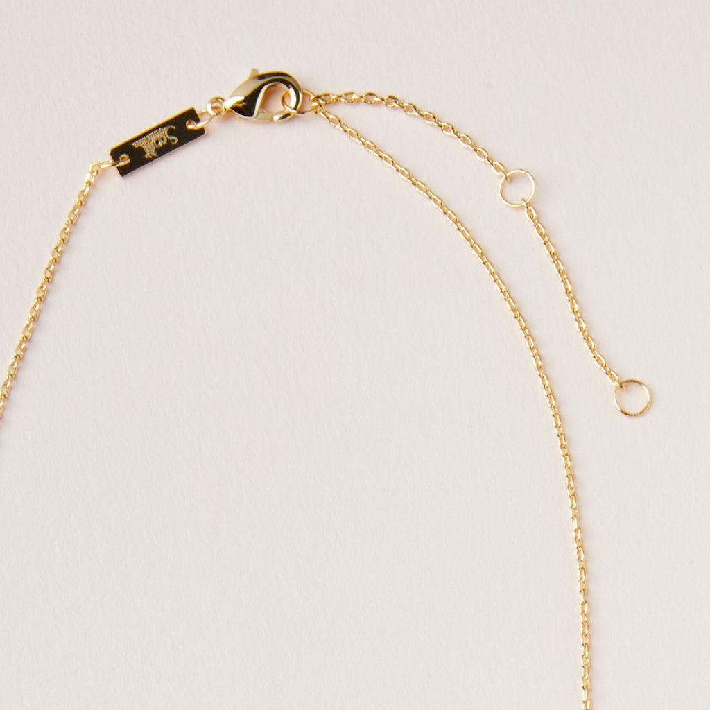 A close-up view of the Scout Jewelry Intention Neck Amethyst/Gold necklace, highlighting its elegant gold chain adorned with a small lobster clasp and an extender chain, all set against a light cream background. The adjustable brass chain is embellished with a tiny rectangular tag near the clasp and includes an engraved charm near the end.