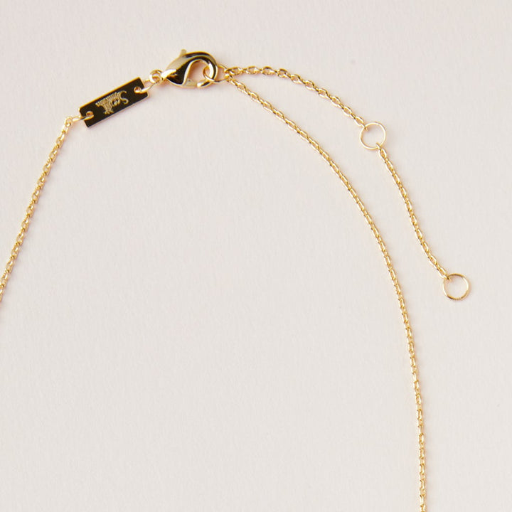A close-up view of the Scout Jewelry Intention Neck Amethyst/Gold necklace, highlighting its elegant gold chain adorned with a small lobster clasp and an extender chain, all set against a light cream background. The adjustable brass chain is embellished with a tiny rectangular tag near the clasp and includes an engraved charm near the end.