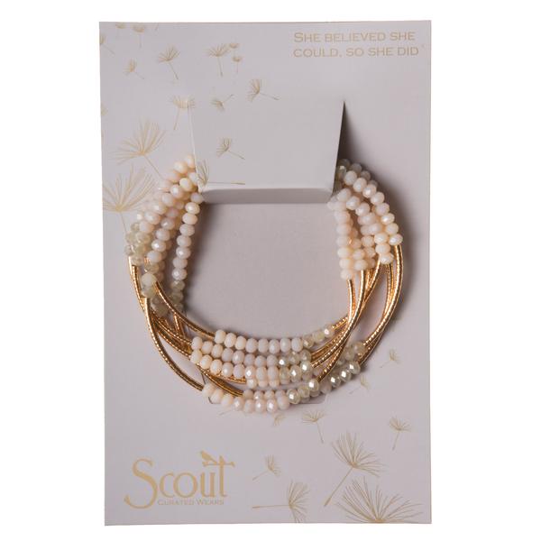 A package showcasing a wrap made of ivory and gold beads, with crystal accents, is presented on a backing card featuring a floral design. The card includes the inspirational text "She believed she could, so she did" and the brand name "Scout Jewelry" at the bottom.