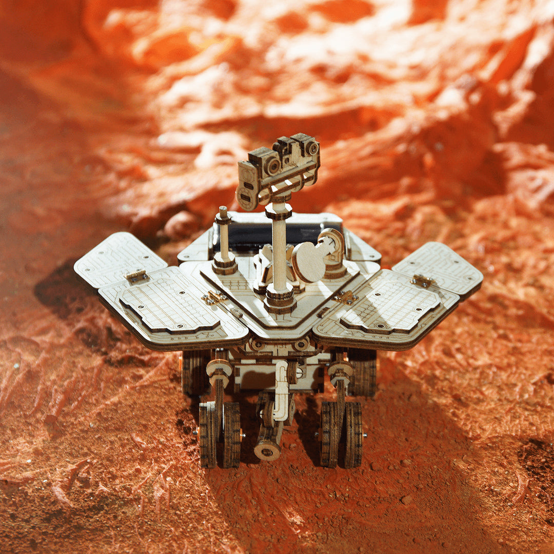 A DIY Solar Powered Rover from Robotime navigates a rocky, reddish surface similar to that of Mars, using its extended panels. Featuring a raised camera mast that casts a shadow as it explores, this model is ideal for educational STEM projects or as an engaging 3D puzzle that vividly brings space exploration to life.