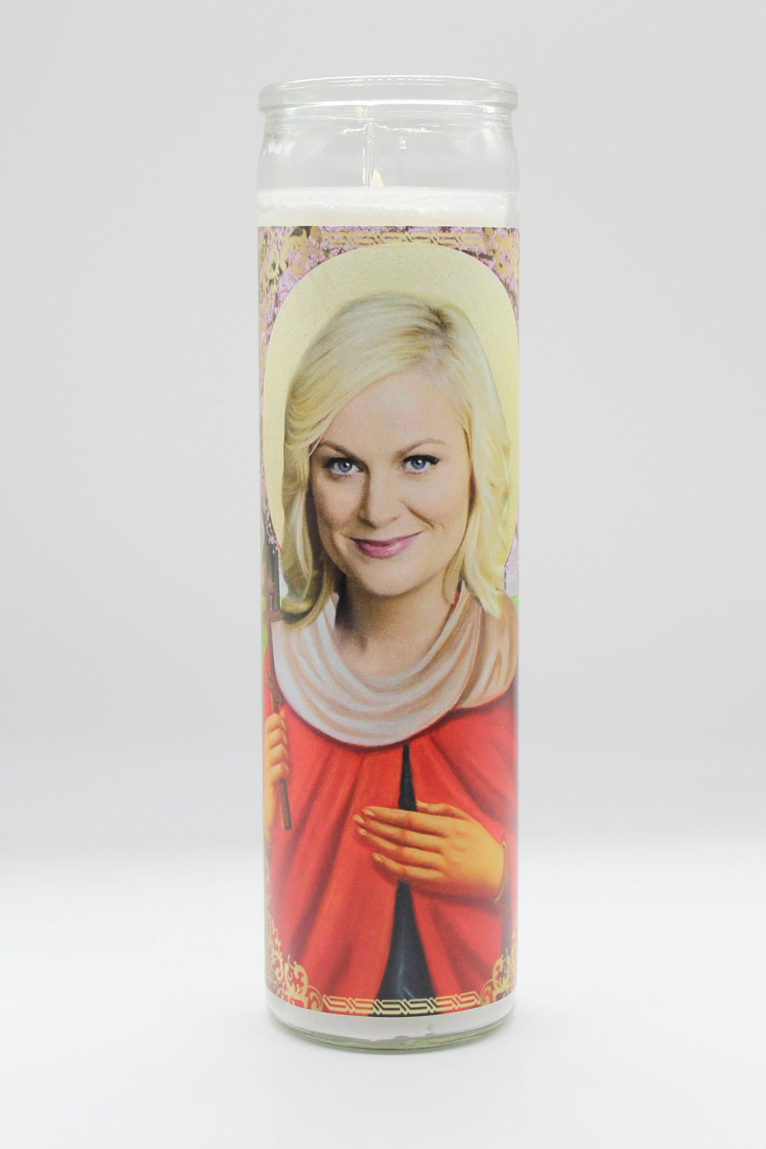 The BOBBYK BOUTIQUE Leslie Knope Candle is a tall white devotional candle showcasing a serene woman in a red robe with blonde hair and holding a scepter, set against a decorative background, reminiscent of a celebrity shrine honoring Lil' Sebastian.